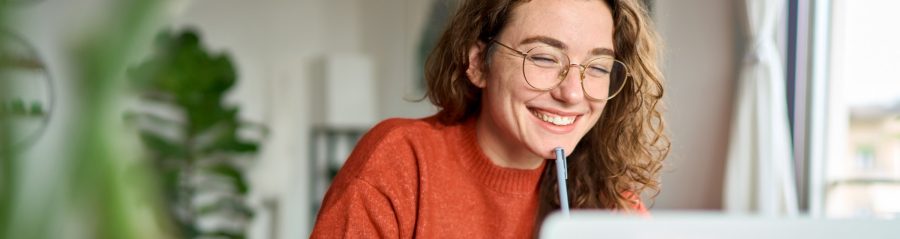 Happy young woman using laptop sitting at desk writing notes while watching webinar, studying online, looking at pc screen learning web classes or having virtual call meeting remote working from home.