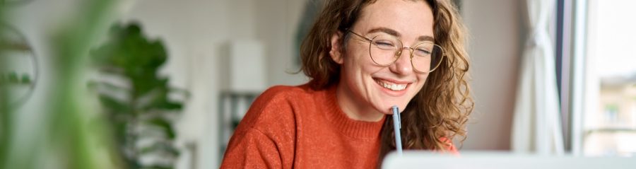 Happy young woman using laptop sitting at desk writing notes while watching webinar, studying online, looking at pc screen learning web classes or having virtual call meeting remote working from home.
