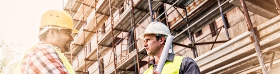 Two men on building site, wearing protective headgear and hi vis jackets