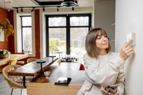 Woman controlling home temperature with electronic thermostat mounted on the wall in living room at home.