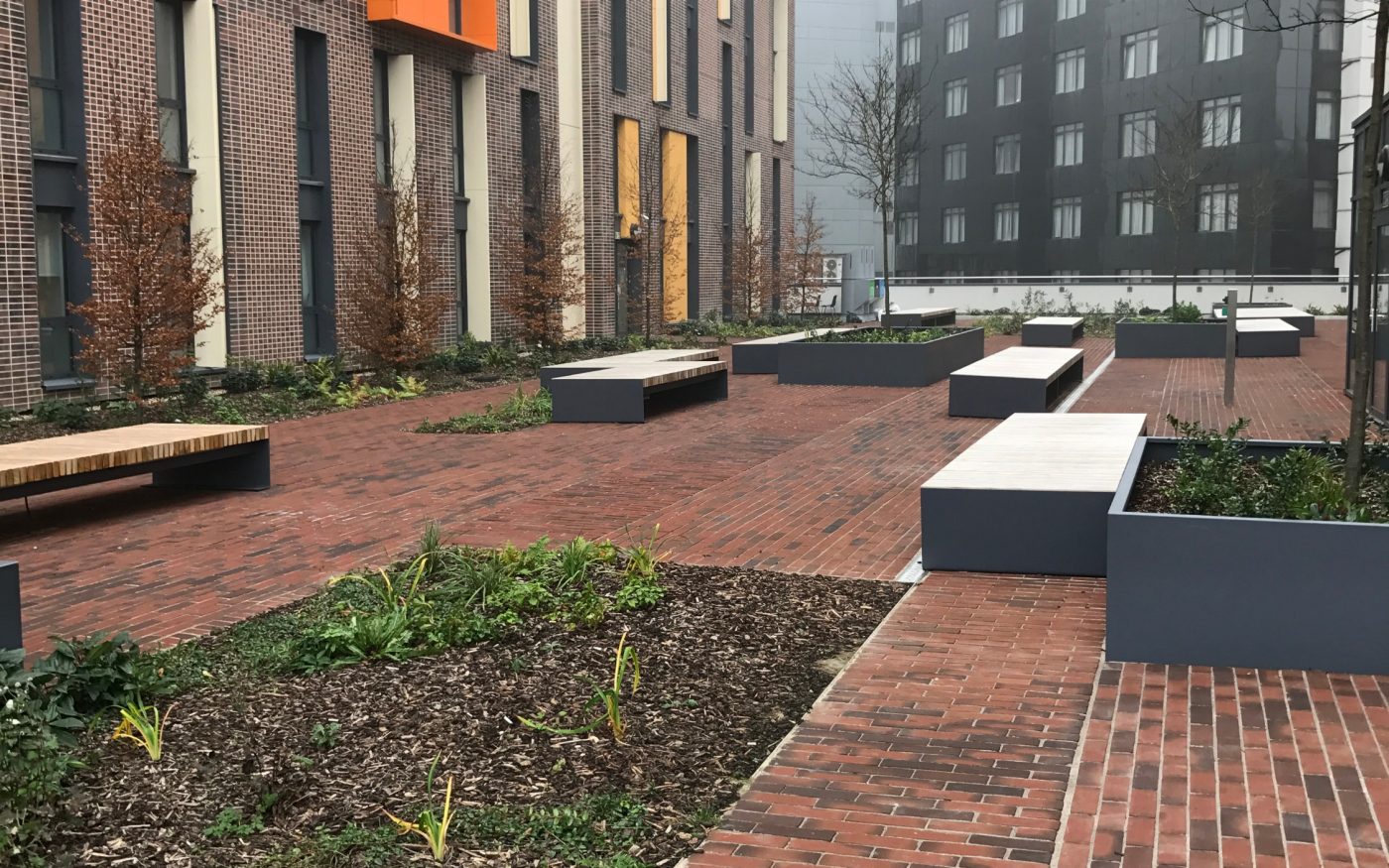 Clay paved path shown in perspective, surrounded by buildings, benches, and greenery