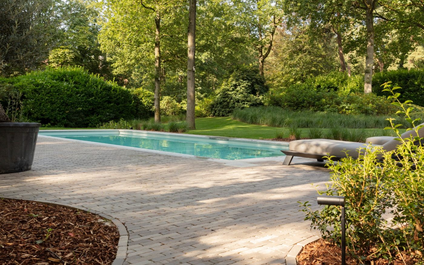 Shot showing paved path surrounding pool, with greenery/trees in background of shot
