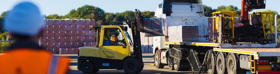 Yellow forklift loading pallet of Wienerberger bricks onto flatbed lorry, with man in high vis vest watching