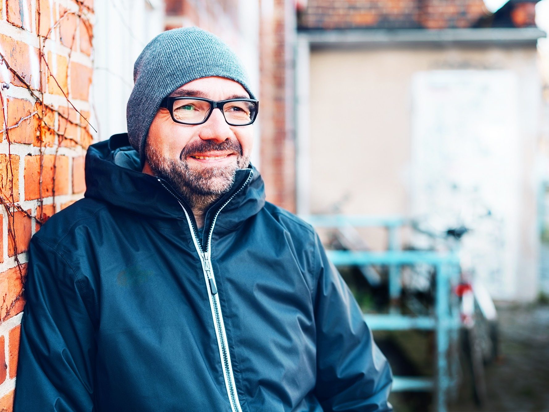Man staring into the distance smiling with grey hat on, leaning against a brick wall