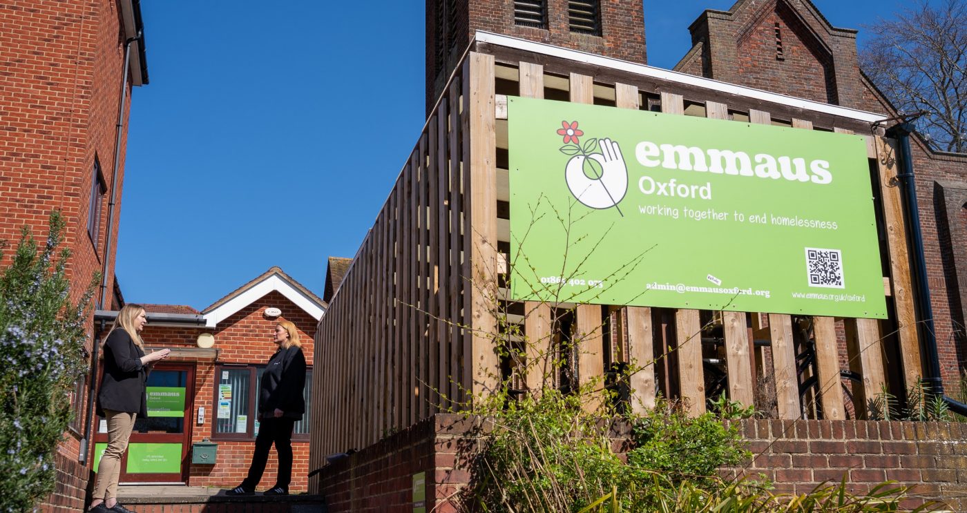 Large green Emmaus sign on the side of a building with two women talking stood on the left