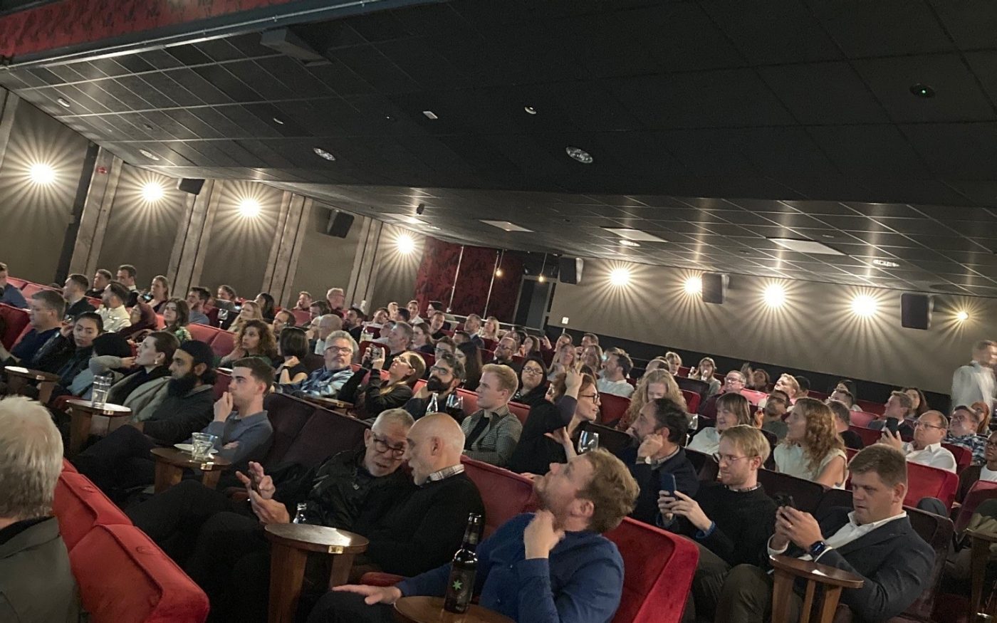 Audience full of people sat on red chairs in theatre