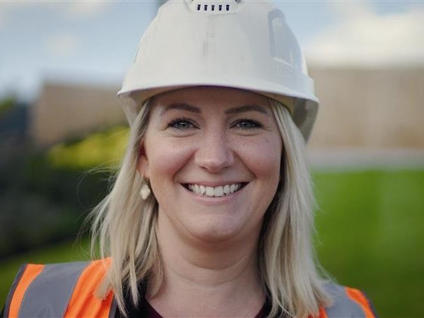 Women smiling towards the camera wearing a white hard hat and orange hi vis jacket