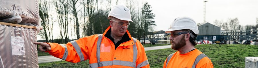 Two men in PPE talking on site, pointing at packaged products outside