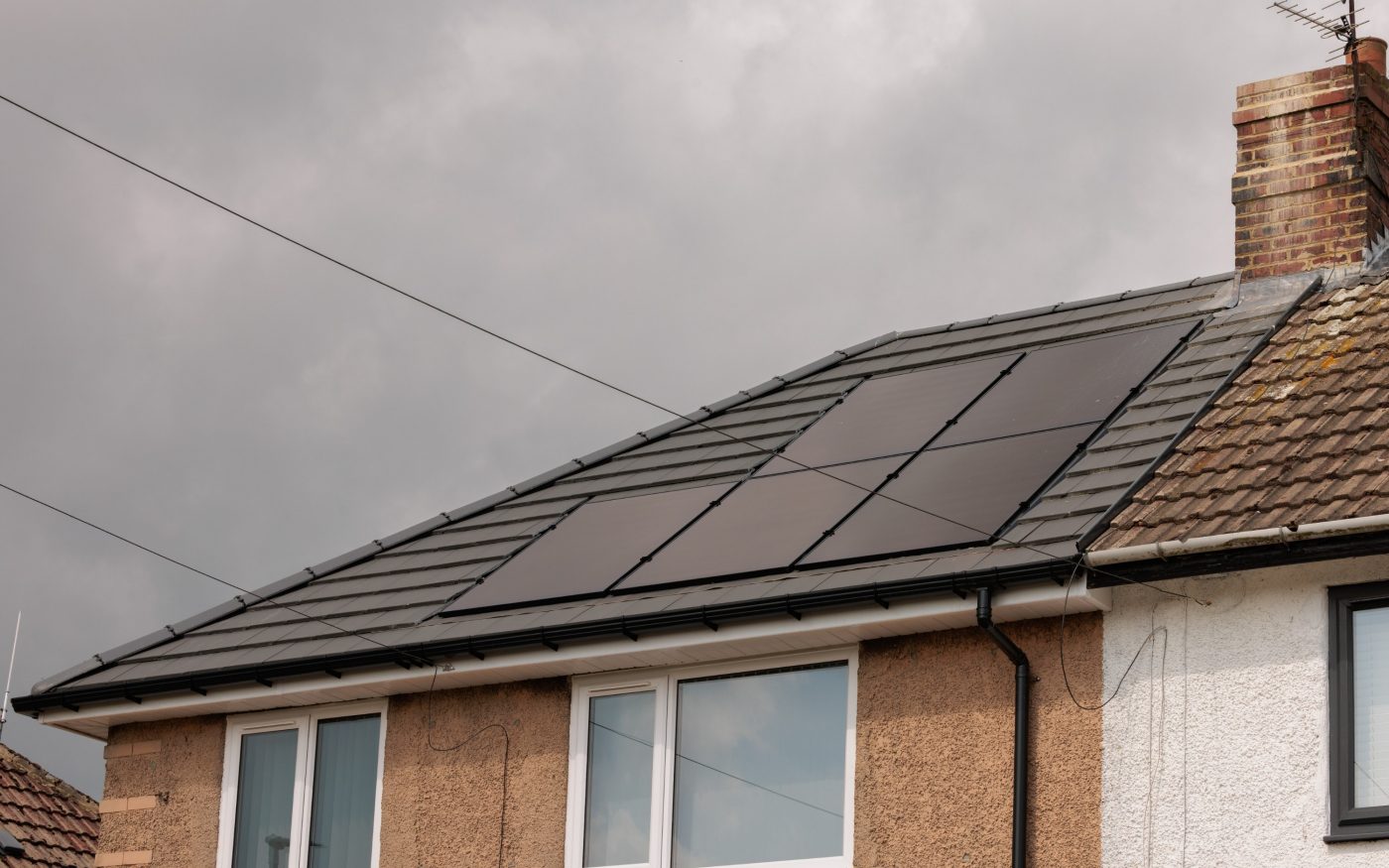 Close-up of a modern house roof with integrated solar panels installed among dark grey tiles under a cloudy sky.